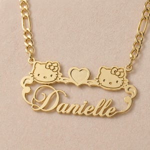 Peut inclure: Un collier doré avec deux motifs Hello Kitty encadrant un cœur. Le nom "Danielle" est écrit en cursive en dessous des motifs Hello Kitty. Le collier est orné d'une chaîne dorée.