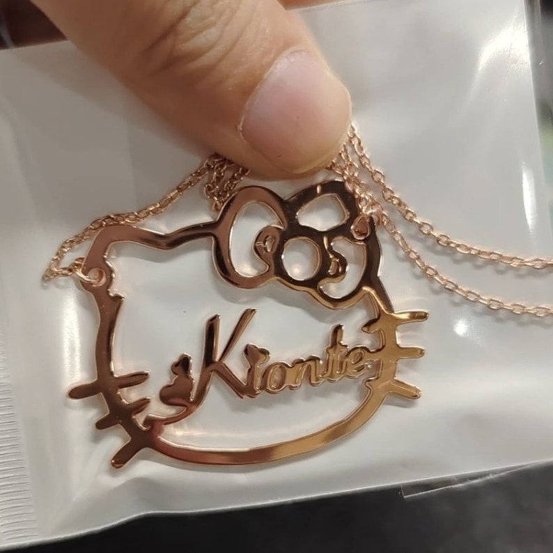 May include: A gold-tone necklace featuring a Hello Kitty charm with the name "Kionte" inscribed within the outline of the character. The necklace is displayed against a white backdrop.