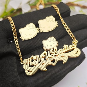 Peut inclure: Ensemble de bijoux dorés comprenant un collier et des boucles d'oreilles assorties. Le collier porte l'inscription "My Ladies" et un motif Hello Kitty. Les boucles d'oreilles sont en forme de Hello Kitty. L'ensemble est présenté sur une surface noire.