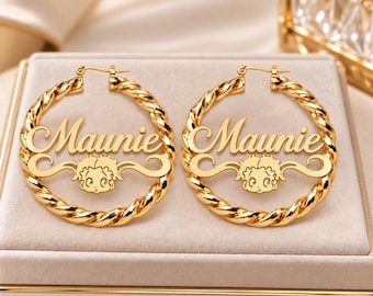 May include: A pair of gold-toned hoop earrings featuring a twisted metal design. Each earring displays the name Maunie in a cursive script font, positioned above a stylised cartoon character face with large eyes and wavy hair.