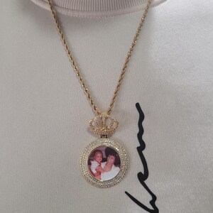 Puede incluir: Collar dorado con un colgante circular que muestra una foto de una persona sosteniendo a un ni&ntilde;o. El colgante est&aacute; rodeado de peque&ntilde;as piedras transparentes y rematado con un dise&ntilde;o de corona. La cadena tiene una textura de cuerda.