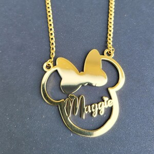 Puede incluir: Collar de oro con un colgante de cabeza de Mickey Mouse con un lazo y el nombre "Maggie" inscrito en el interior.