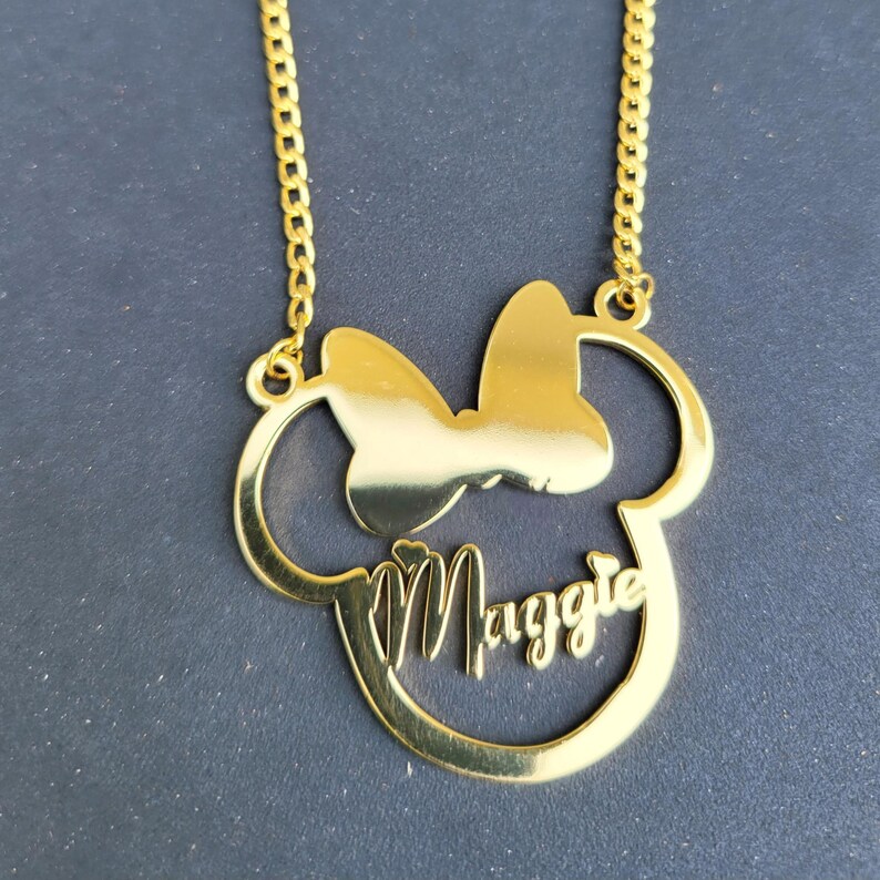 Puede incluir: Collar de oro con un colgante de cabeza de Mickey Mouse con un lazo y el nombre "Maggie" inscrito en el interior.
