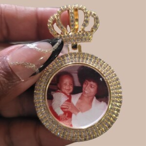 Puede incluir: Un colgante dorado con un dise&ntilde;o de corona en la parte superior. El colgante tiene un marco circular incrustado con peque&ntilde;as piedras transparentes. Dentro del marco hay una fotograf&iacute;a de una mujer y un ni&ntilde;o. La corona tambi&eacute;n est&aacute; adornada con piedras transparentes.
