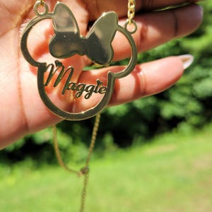 Puede incluir: Collar de tono dorado con un colgante de cabeza de Mickey Mouse con un lazo y el nombre "Maggie" inscrito en el interior.