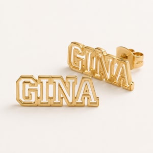 May include: A pair of gold-toned stud earrings featuring the name GINA in a block letter varsity font. The letters have a hollow, open-frame design with a metallic gold finish and a post-back closure mechanism visible on one of the earrings.