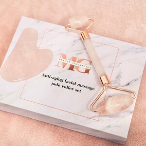 May include: A white box with a marble pattern and gold lettering that says "Beauty Collection MG" and "Anti-aging facial massage jade roller set". The box contains a pink jade roller and a pink jade gua sha tool.