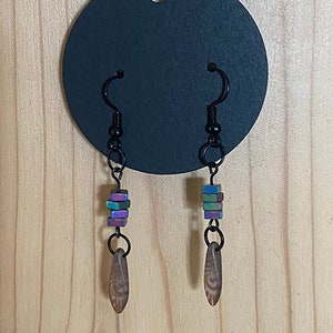 May include: A pair of dangle earrings with iridescent purple, blue, and green beads and a brown wood-like charm. The earrings are hanging from a black card.