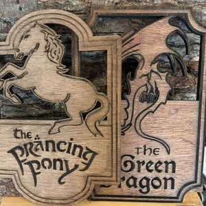 May include: Wooden wall art with two signs. One shows a rearing horse with the text "The Prancing Pony." The other depicts a dragon with the text "The Green Dragon." Both signs are made of wood with a dark border.