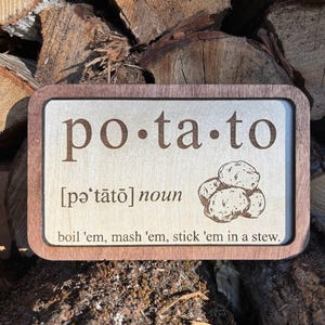 Könnte beinhalten: Rechteckiges Holzschild mit hellem Holzmaserungszentrum und dunkelbraunem Rand. Das Wort "potato" ist in einer Serifenschrift eingraviert, zusammen mit einer Aussprachehilfe und einer Zeichnung von Kartoffeln. Der Text "boil 'em, mash 'em, stick 'em in a stew" ist ebenfalls eingraviert.
