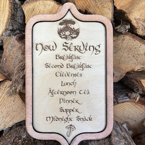 May include: Wooden sign with a light brown frame and a cream-coloured centre. The sign reads "Now Serving" and lists meal times in a stylized font. A circular graphic of a hobbit hole is at the top. The sign is set against a backdrop of stacked firewood.