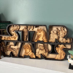 May include: Wooden Star Wars sign with laser-cut characters and the Star Wars logo. The sign is dark brown with light brown character cutouts. A Lego Baby Yoda figure is on the right.