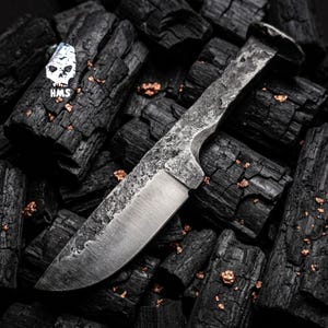 May include: A handcrafted, rustic knife with a textured, gray metal blade and handle. The knife rests on a bed of dark charcoal. The handle has a unique shape, and the blade is sharp. The image includes a skull logo with the letters "HMS".