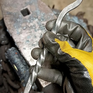 May include: A hand-forged metal hook with a twisted handle and a curved end. The hook is held by a gloved hand, with an anvil in the background. The HMS logo is visible in the upper left corner.