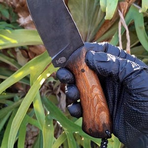 Peut inclure: Un couteau à lame noire avec un manche en bois. Le couteau porte un logo de crâne sur la lame et une lanière. Le manche est de couleur marron clair avec un grain de bois. Le couteau est tenu par une personne portant un gant noir.