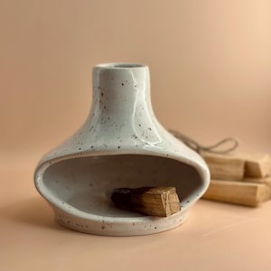 May include: A speckled white ceramic incense holder with an oval opening, containing a piece of Palo Santo wood. The holder has a rounded shape and a small opening at the top. A bundle of Palo Santo sticks is in the background.