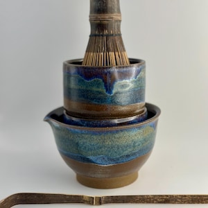 Puede incluir: Un juego de té matcha de cerámica con un batidor de bambú, un cuenco y una cuchara. El cuenco y el soporte del batidor están esmaltados con tonos azules y marrones. El batidor de bambú es marrón oscuro.