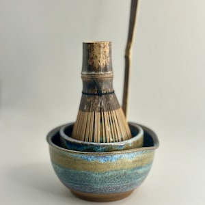 May include: A brown and blue ceramic matcha bowl with a bamboo whisk and a wooden whisk holder.