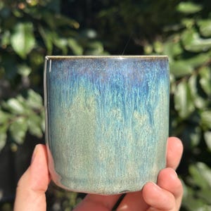 Puede incluir: Una taza pequeña de cerámica con un esmalte azul y verde. El esmalte tiene un aspecto moteado y texturizado.