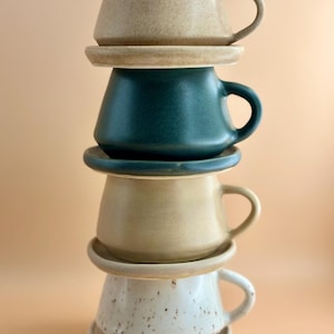 May include: Four ceramic coffee cups and saucers are stacked. The cups are in beige, teal, tan, and speckled white. Each cup has a rounded handle. The cups are stacked, forming a tower. The saucers are round.