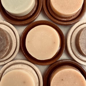 May include: Overhead shot of several round soaps in various shades of beige and brown, each resting on a matching ceramic soap dish. The soaps have a smooth, slightly textured surface. The arrangement is symmetrical, showcasing the handmade quality.