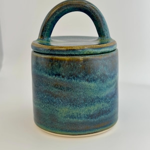 May include: A ceramic jar with a lid and handle. The jar is cylindrical with a dark teal and blue glaze. The handle is arched and the lid fits snugly on top. The base of the jar is a light cream colour.