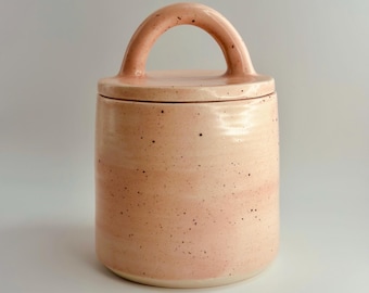May include: A peach-coloured ceramic jar with a lid and handle. The cylindrical jar has a speckled finish and a curved handle on the lid. The design is minimalist and modern, suitable for kitchen or home decor.