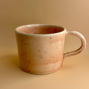 May include: A peach-coloured ceramic mug with a speckled glaze. The mug has a rounded body, a curved handle, and a white interior. The mug is a unique piece of pottery.