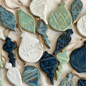 May include: A selection of ceramic Christmas tree decorations in shades of blue, white, and green. Each ornament has a unique design and is strung with twine. The decorations are arranged on a textured, off-white surface.