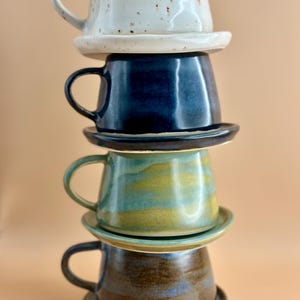 May include: Four ceramic mugs and saucers stacked on top of each other. The mugs are in white, navy blue, teal, and brown. Each mug has a handle and a flared shape. The saucers are round and match the mugs. The mugs are made of ceramic.