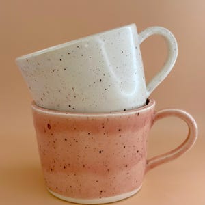 May include: Two ceramic mugs, one stacked on the other. The bottom mug is pink with speckles, the top one is white with speckles. Both have curved handles and a smooth, glossy finish. The mugs are set against a peach-coloured background.