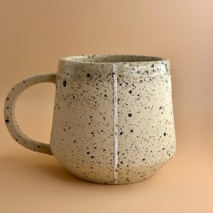 May include: A speckled ceramic mug with a rounded base and a large handle. The mug is off-white with black speckles and a vertical white line. The mug is designed for hot beverages.