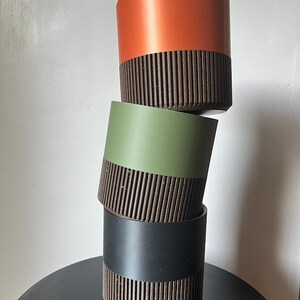 May include: Three stacked plant pots, the top one is orange, the middle one is green, and the bottom one is black. Each pot has a brown ribbed band around the middle.