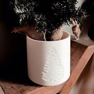 May include: A white ceramic planter with a debossed Christmas tree design. The planter holds a small artificial evergreen tree wrapped in burlap. The planter sits on a wooden shelf, creating a festive holiday display.