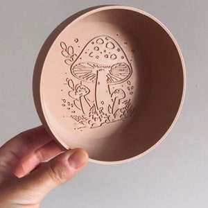 May include: A small, round, peach-coloured ceramic bowl with an etched design of mushrooms and foliage. The bowl is held in a hand, showcasing the intricate artwork. The design features a large mushroom with smaller ones around it.