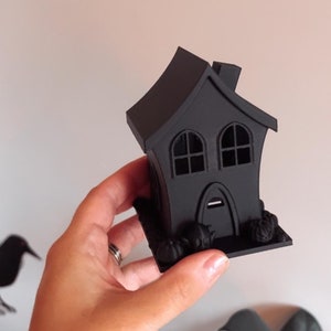 Op de afbeelding: Een zwart 3D-geprint Halloween-huis met twee ramen en een deur. Het huis staat op een basis met kleine pompoenen eromheen.
