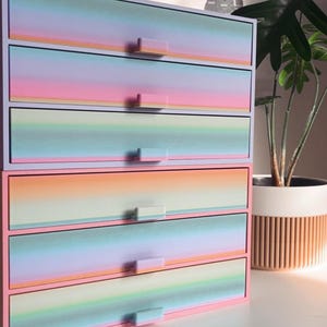 May include: A five-drawer storage chest in pastel colours. The drawers have a gradient design with pink, blue, and yellow hues. Each drawer has a small, rectangular handle. The chest is set against a backdrop of a world map and a potted plant.