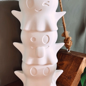 May include: Three white ceramic ghost planters stacked on top of each other. The top ghost has a winking face, the middle ghost has a tongue sticking out, and the bottom ghost has two eyes.