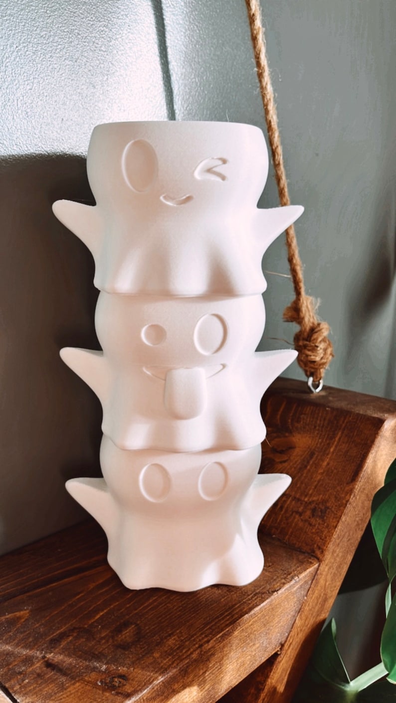 May include: Three white ceramic ghost planters stacked on top of each other. The top ghost has a winking face, the middle ghost has a tongue sticking out, and the bottom ghost has two eyes.