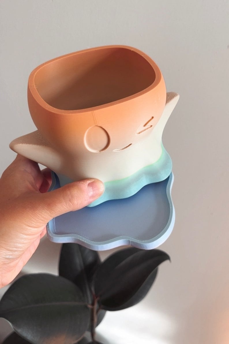 May include: A pastel-colored ghost-shaped planter with a blue and white ombre design. The planter has a square opening at the top and a scalloped base. The planter is sitting on a matching blue scalloped saucer.