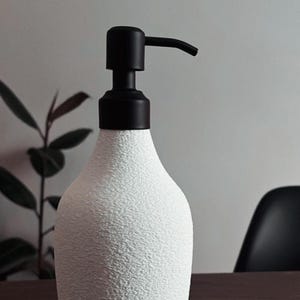 May include: White ceramic soap dispenser with a black pump. The dispenser has a textured surface.