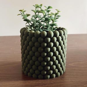 May include: A cylindrical planter with a textured exterior composed of numerous small, matte olive green spheres arranged in a grid pattern. A small, artificial green plant with multiple stems and oval-shaped leaves sits inside the container.
