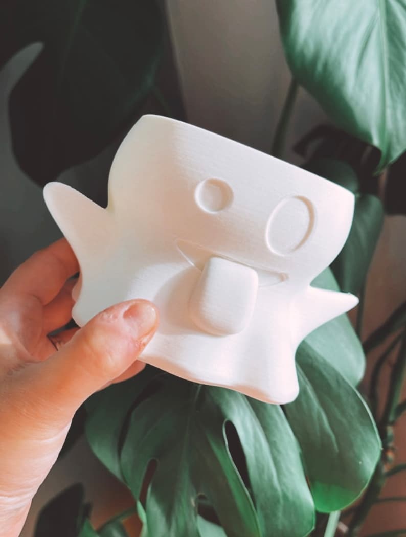 May include: A white, ghost-shaped planter with a smiling face and a protruding tongue. The planter has a rounded body with two small arms and is held in a hand. The background features large green leaves.