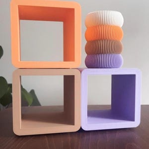 May include: A selection of colourful, geometric home decor items. The image shows four square, open-sided boxes in shades of orange, tan, and purple. A stack of ribbed, cylindrical objects in white, orange, brown, and purple sits atop one of the boxes.