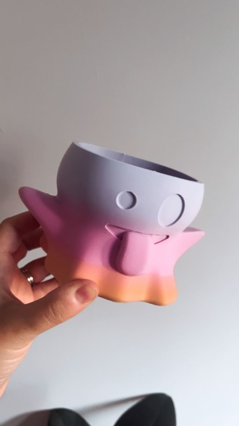 May include: A whimsical, ghost-shaped planter in a gradient of colors. The top is lavender, transitioning to pink and then orange at the base. It features two circular eye cutouts and a small, rectangular tongue.