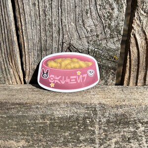 Puede incluir: Una pegatina rosa que muestra un cuenco lleno de nuggets dorados. El cuenco está decorado con un icono de conejo en blanco y negro, un icono de calavera blanca, dos estrellas amarillas y texto rosa estilizado que dice OKLIVIN7 al frente.