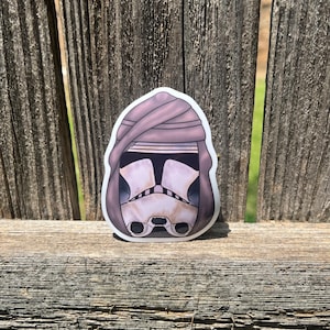 Puede incluir: Una pegatina troquelada que muestra un casco de soldado clon blanco con detalles de visera negros, parcialmente cubierto por una capucha de tela marrón claro drapeada. La ilustración tiene un borde blanco limpio y un acabado mate suave.