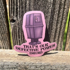 Puede incluir: Una pegatina troquelada que muestra un droide de energía morado y cuadrado con pies pequeños y un panel de control con luces rojas y amarillas. La pegatina tiene un fondo rosa claro y el texto negro, THAT'S OUR DEFECTIVE POWER DROID.