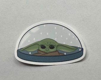 May include: A die-cut sticker featuring a green, large-eared creature with dark eyes wrapped in a brown cloak, enclosed within a clear, dome-shaped snow globe with a dark blue base and small white dots representing falling snow inside the dome.