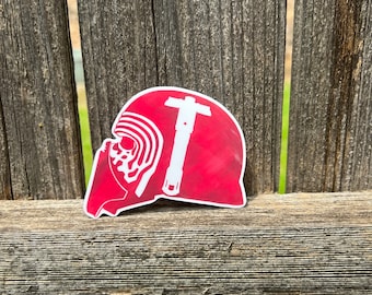 May include: A die-cut sticker featuring a red background with a white silhouette of a helmeted figure and a cross-guard lightsaber. The design displays a textured, painted appearance with a white border outlining the entire shape of the sticker.